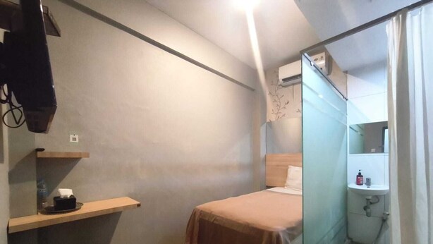 A small, compact room with a bed, a stool, a shelf with a bottle, and a bathroom with a sink and mirror. - Collection O near Lampung City Mall formerly Royal Guesthouse (Bandar Lampung)