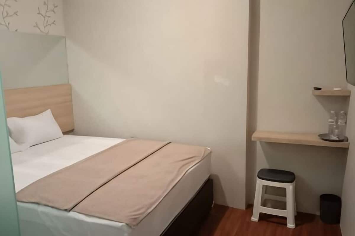 Un dormitorio pequeño y ordenado con cama, un taburete, una estantería con objetos y un cubo de basura.