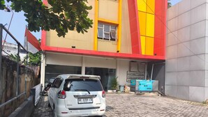 A white car parked in front of a building with a red and yellow facade, a paved ground, and a visible signboard. - Collection O near Lampung City Mall formerly Royal Guesthouse (Bandar Lampung)