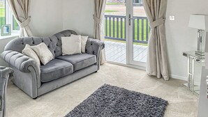 A grey sofa with white cushions, a grey rug, a lamp, and a view of a deck and greenery through large windows. - 2 bedroom accommodation in Laurenkirk (Laurenkirk)