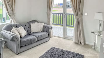 Un canapé gris avec des coussins blancs, un tapis gris, une lampe et une vue sur une terrasse et la verdure, le tout visible à travers de grandes fenêtres.