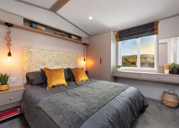 A modern bedroom with a bed, bedside tables, a headboard with a textured pattern, a window with a view, and built-in shelves. - A recent addition to the Searles Leisure Resort holiday hire fleet. (Hunstanton)