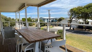 A covered outdoor area with a wooden table and white chairs, overlooking a residential street with houses and trees.