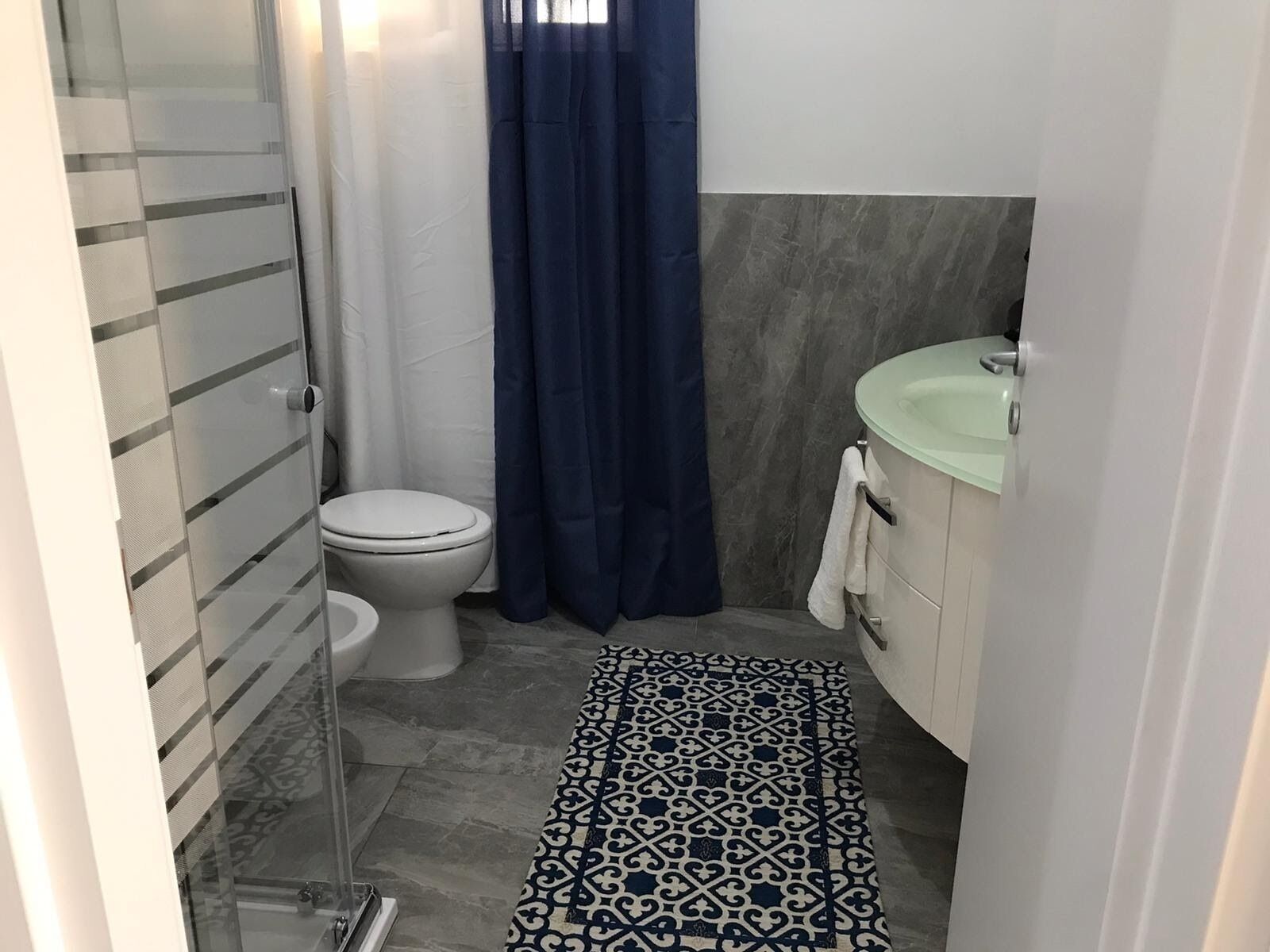 Une salle de bain avec une douche à parois de verre, des toilettes blanches, un rideau de douche bleu, un tapis à motifs et un meuble-lavabo blanc avec un lavabo vert.
