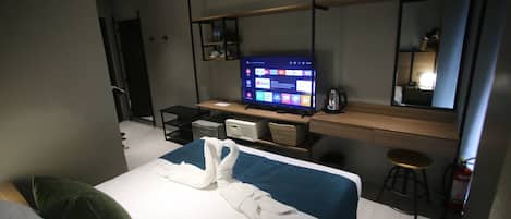A modern hotel room with a bed, a desk with a computer, a TV displaying a digital interface, a mirror, and a fire extinguisher.