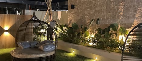 Un espacio al aire libre con asientos nocturnos que incluye una silla reclinable de mimbre, una cama pequeña y una palmera.