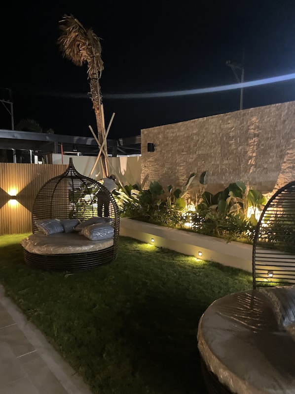 Un espacio al aire libre con asientos nocturnos que incluye una silla reclinable de mimbre, una cama pequeña y una palmera.