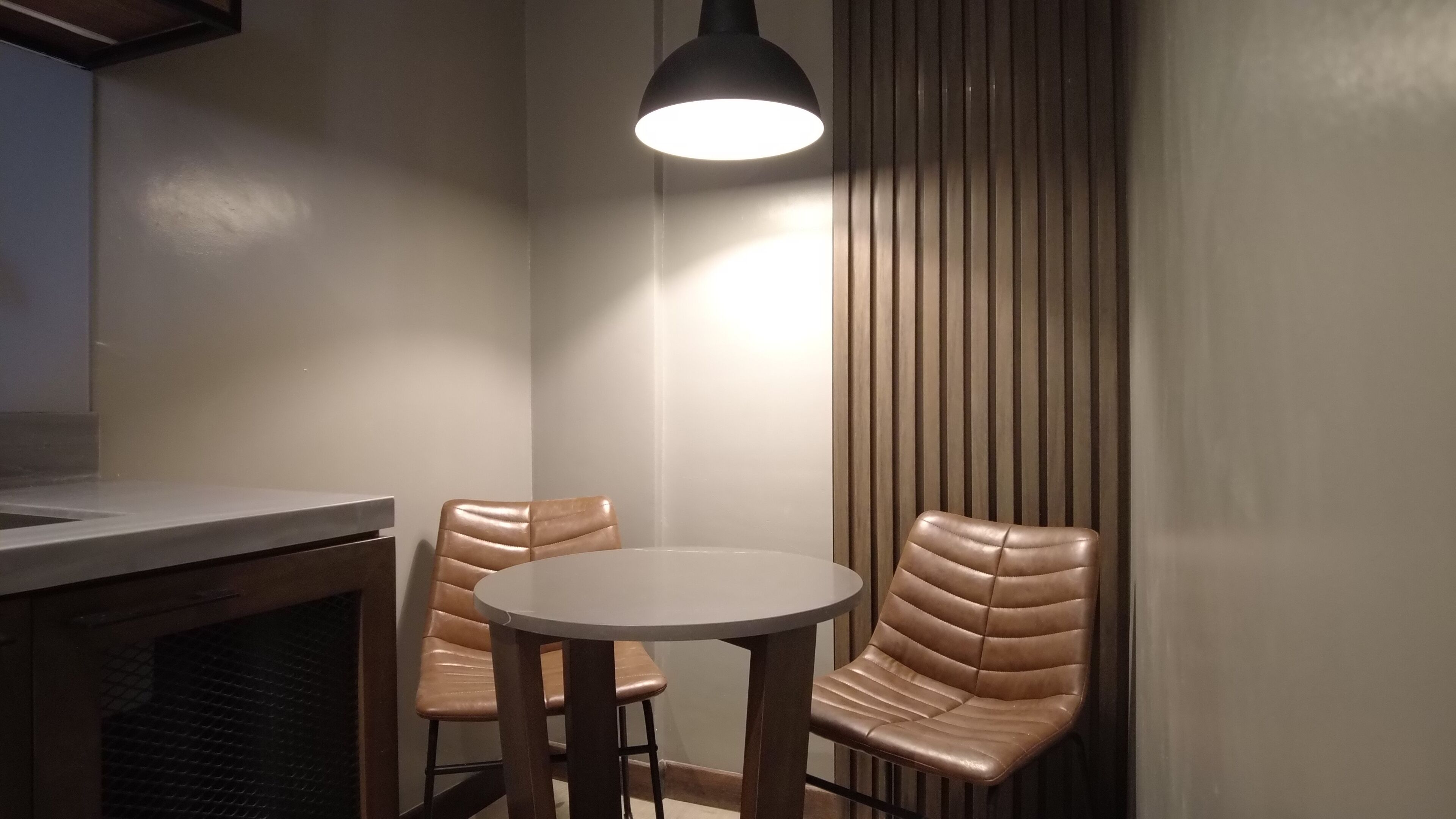 Un pequeño comedor con una mesa redonda y dos sillas de cuero, una lámpara colgante arriba y un panel de pared de listones de madera.