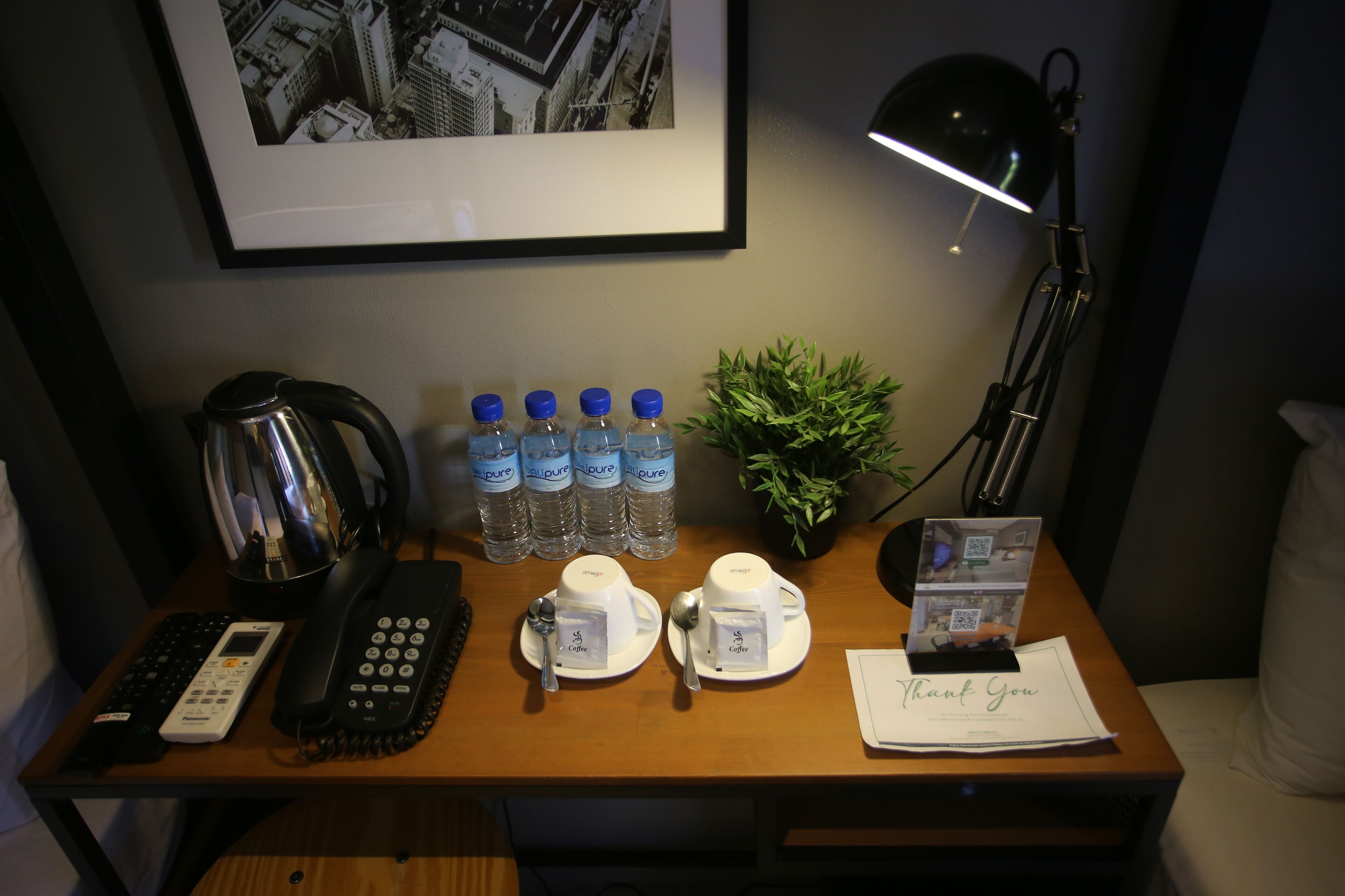 A hotel desk with a telephone, two cups labeled "Coffee," water bottles, a plant, a lamp, and a "Thank You" card.