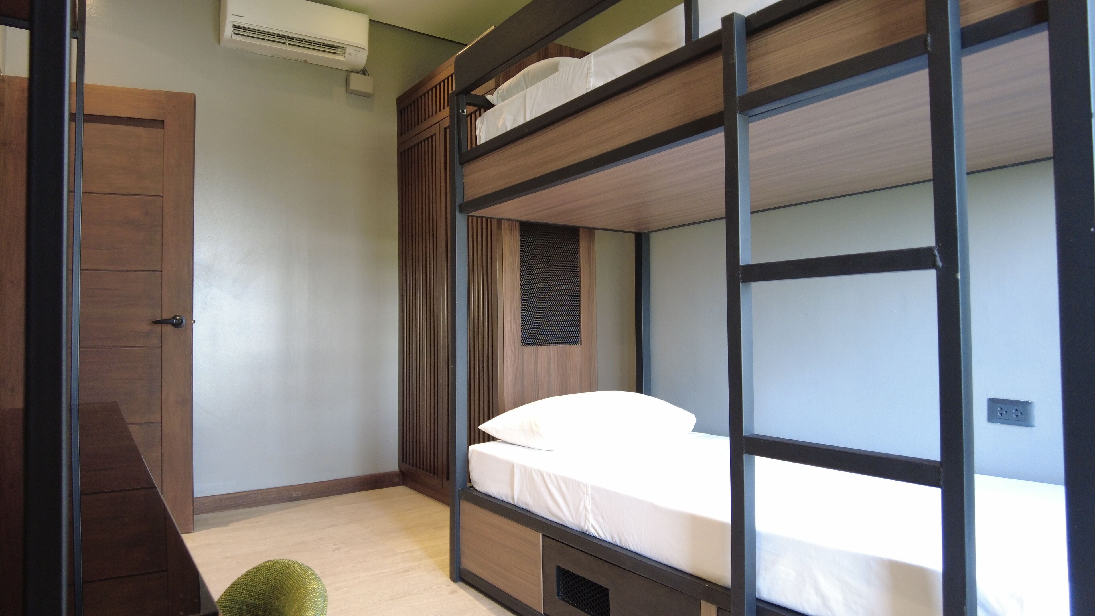 Una cama litera con estructura de madera, un cajón pequeño para guardas y un panel de malla en la pared.