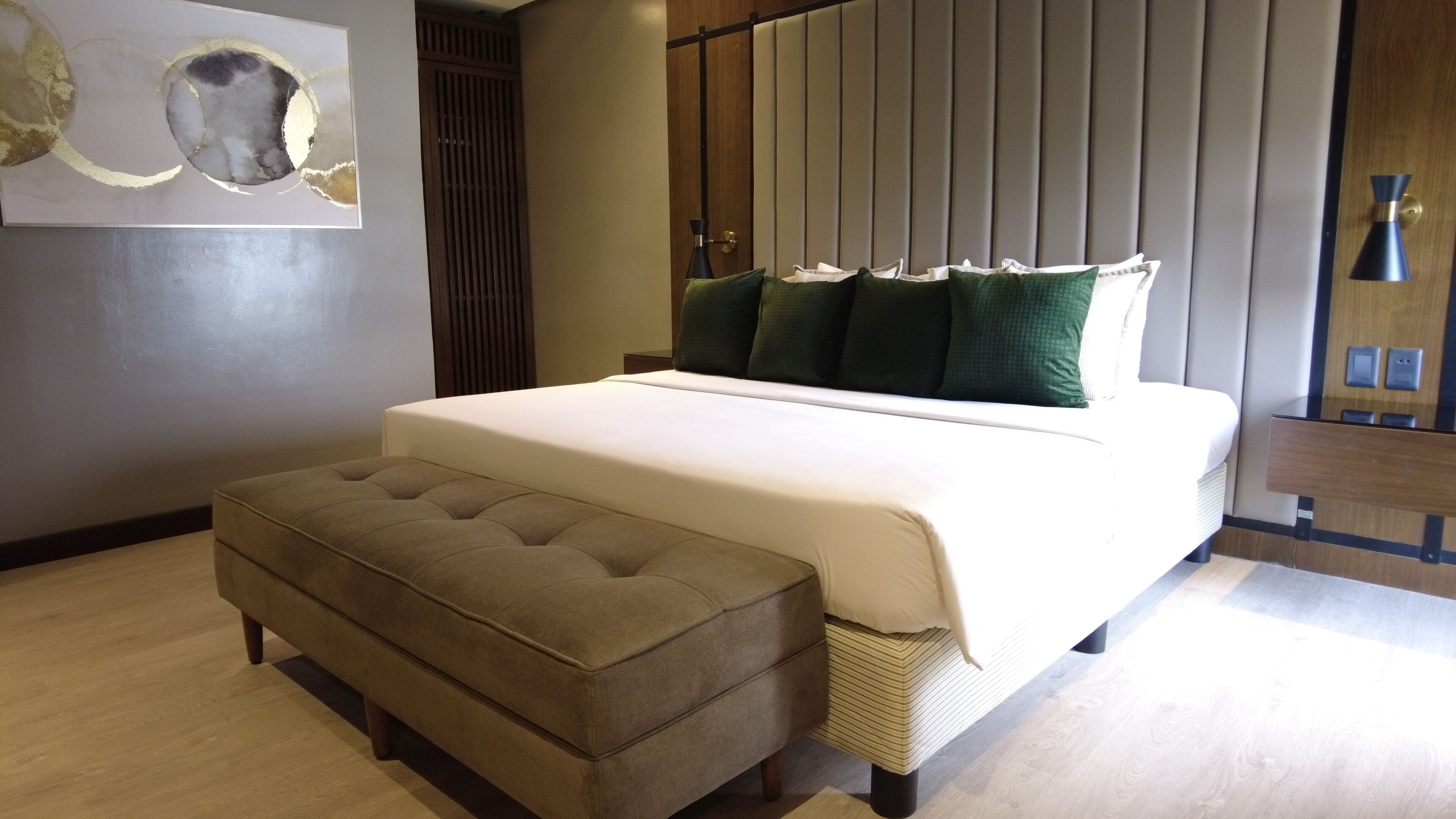 A modern bedroom with a large bed, a tufted ottoman, and a wall-mounted artwork.