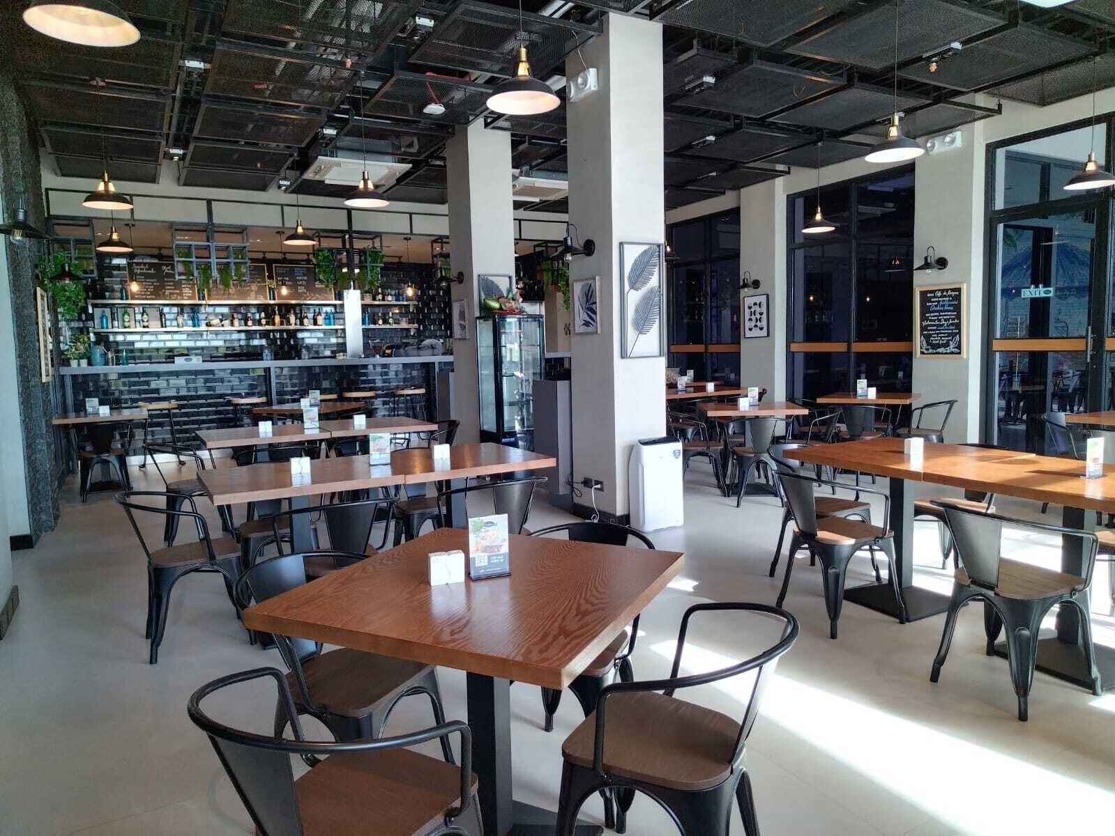 Una cafeteria moderna amb taules de fusta i caires negres, grans finestres i una zona de bar amb prestatges plens de botelles.