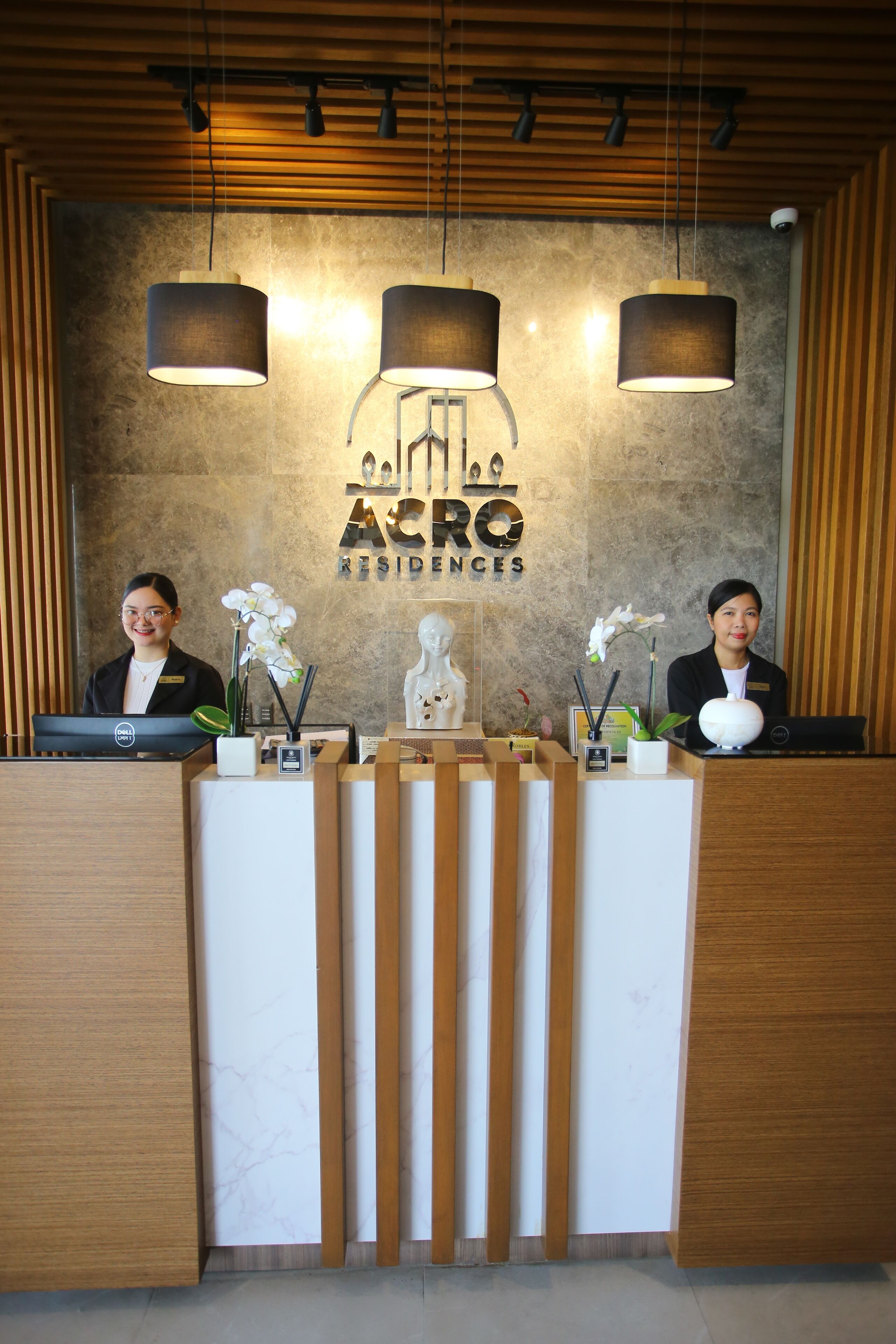 Una recepción con dos empleados, un mostrador de madera, una pared de mármol y el logotipo de "ACRO Residences".