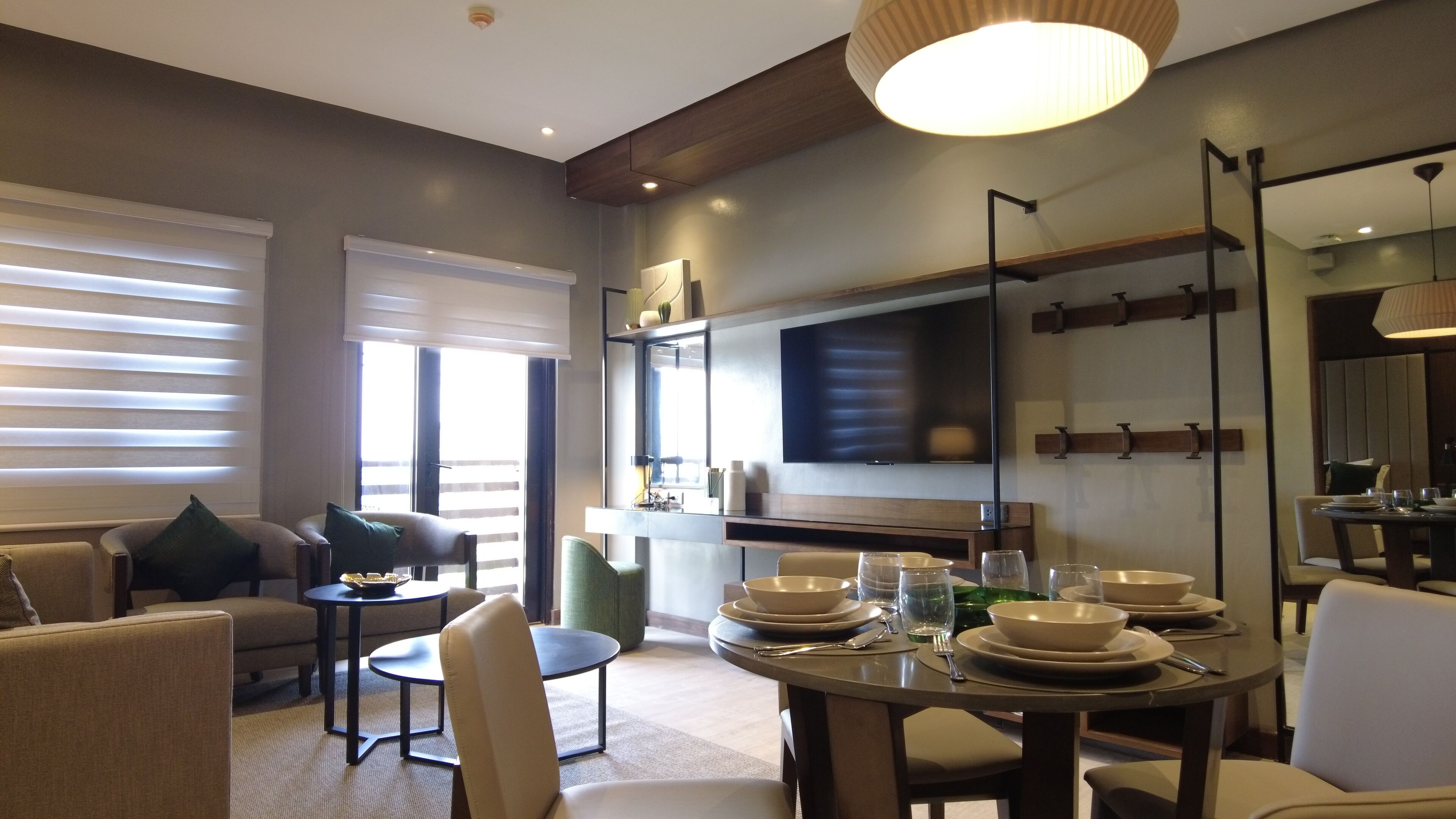 Una sala de estar moderna con zona de comedor, que incluye un televisor de pantalla plana, un amplio ventanal con persianas y una mesa de comedor servida con platos y vasos.