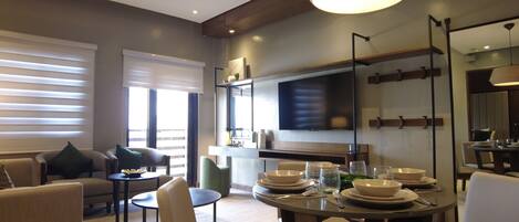 Una sala de estar moderna con zona de comedor, que incluye un televisor de pantalla plana, un amplio ventanal con persianas y una mesa de comedor servida con platos y vasos.