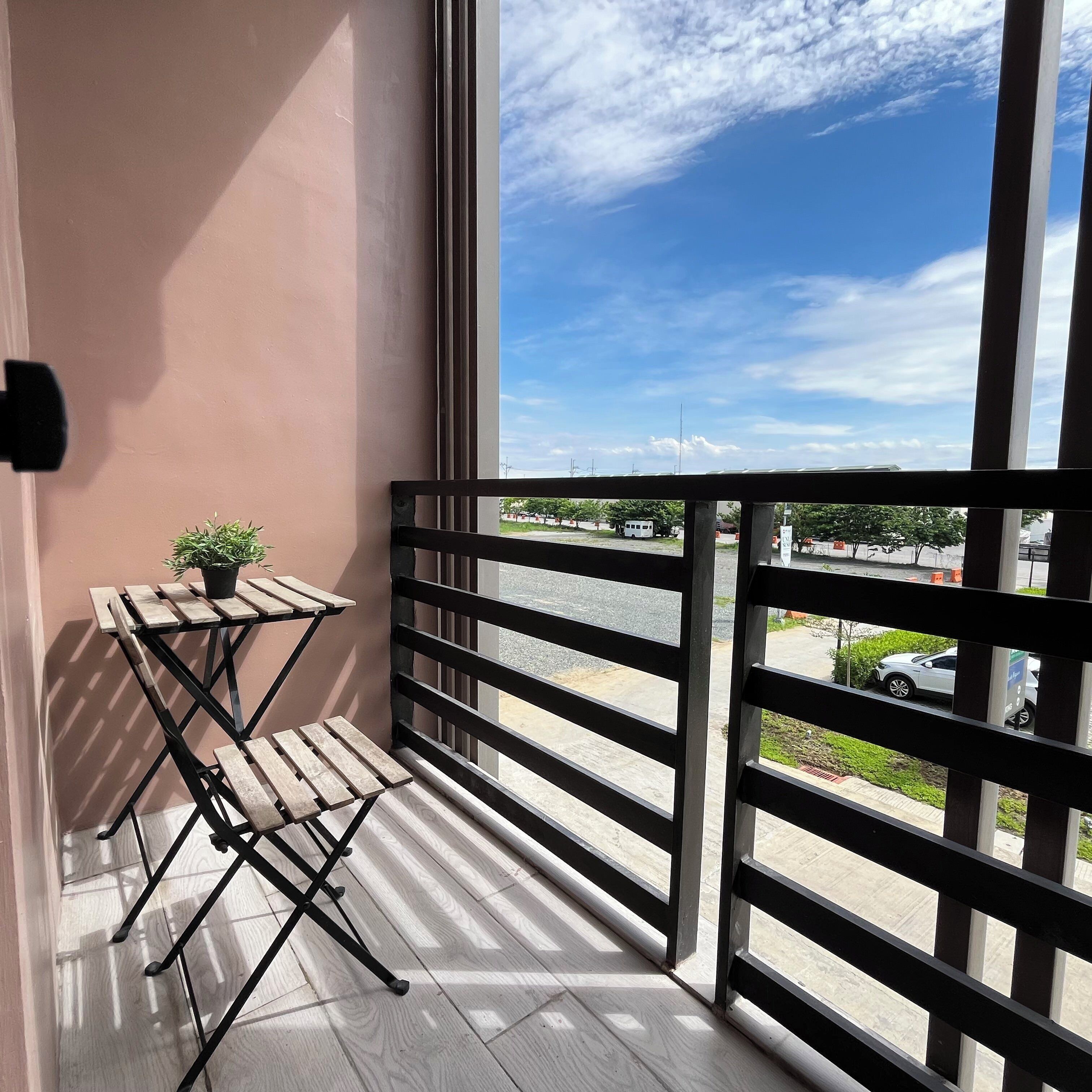 A balcony with a wooden table and chairs, a potted plant, and a view of the street and buildings.