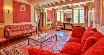Una habitación con un sofá rojo estampado, una mesa de centro de vidrio y un sillón rojo. Hay un espejo en la pared y una puerta que da a otra habitación.