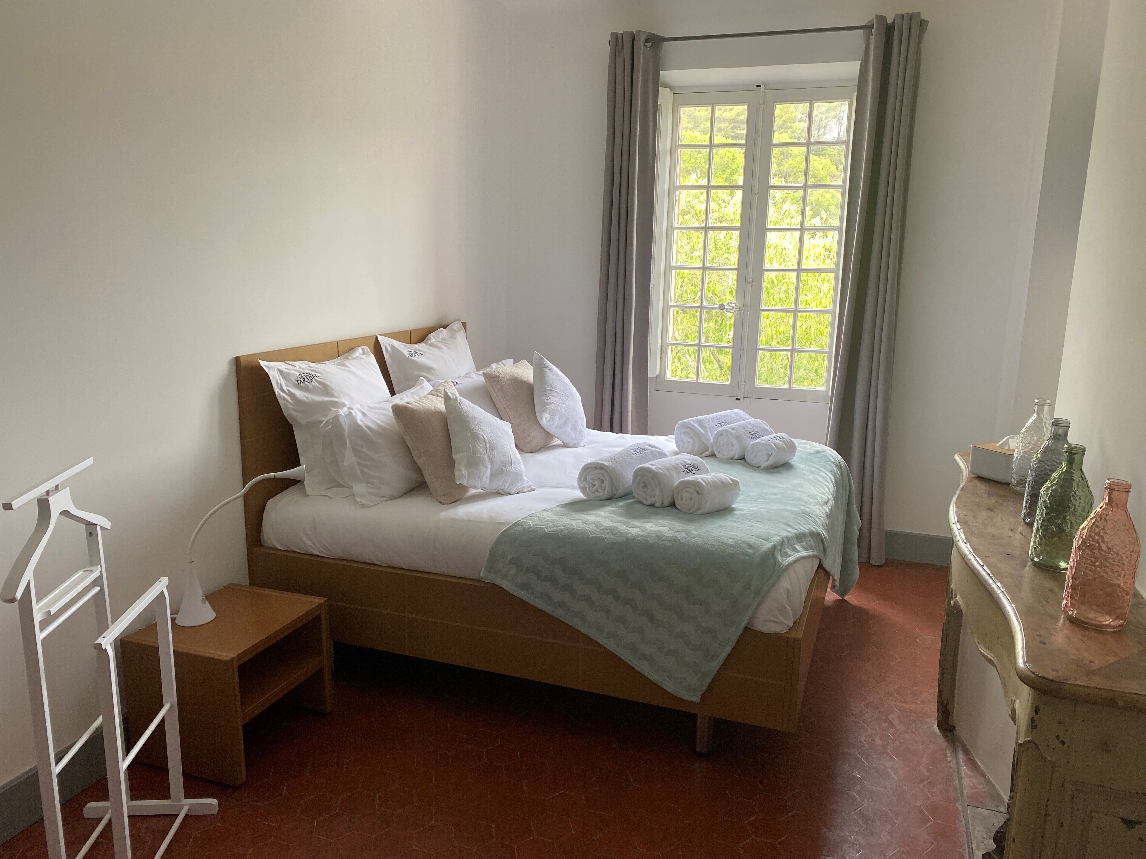A neatly made bed with white linens and pillows, a bedside table with a lamp, and a window with curtains.
