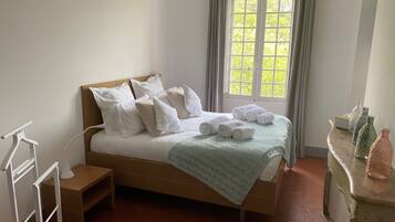 A neatly made bed with white linens and pillows, a bedside table with a lamp, and a window with curtains.