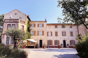 A historic building with a courtyard, featuring a central entrance, multiple windows with shutters, and surrounding greenery.