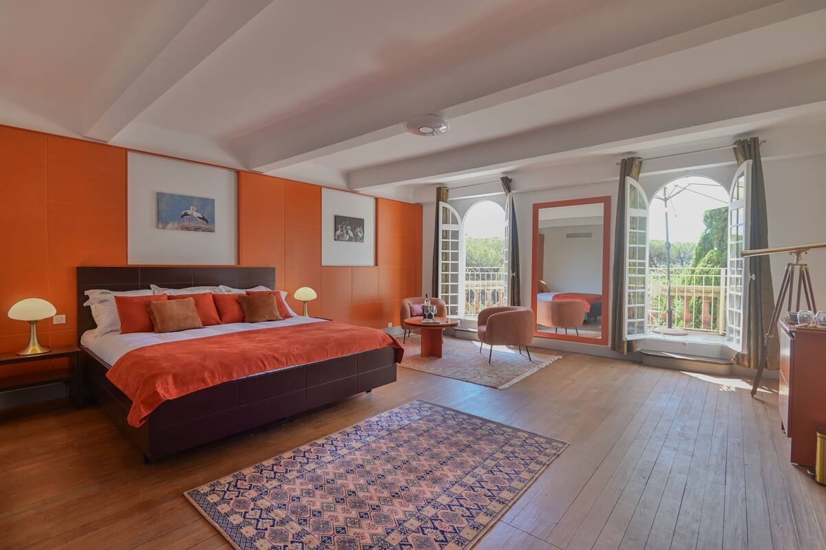 A bedroom with a large bed, a small table, a chair, and a balcony with a view.