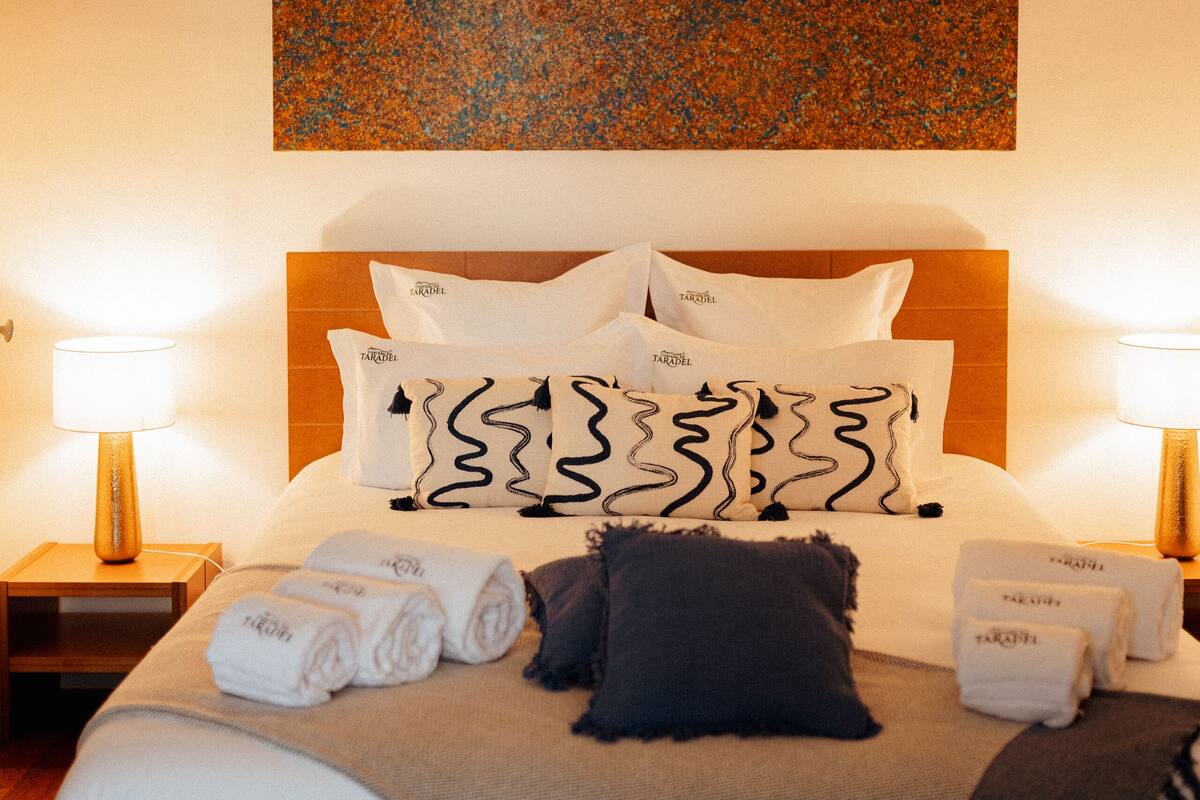 A neatly made bed with white and patterned pillows, a dark throw blanket, and neatly folded towels. There is a wooden headboard and a wall art piece above the bed.