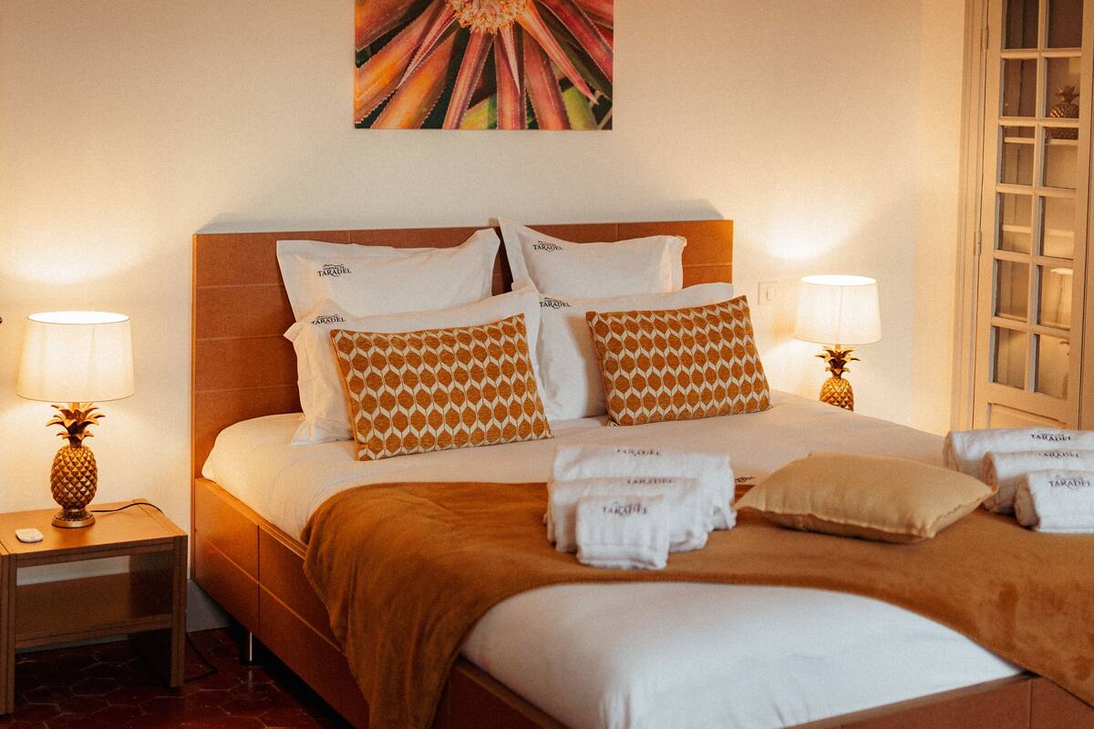 A hotel room with a bed, two bedside tables with lamps, a pineapple on one table, and a large floral artwork on the wall.