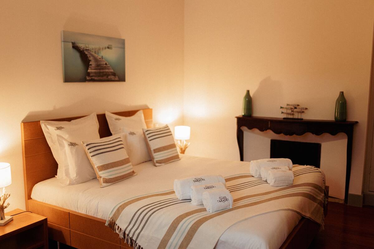 A neatly made bed with striped bedding and pillows, a bedside table with a lamp, and a painting on the wall.