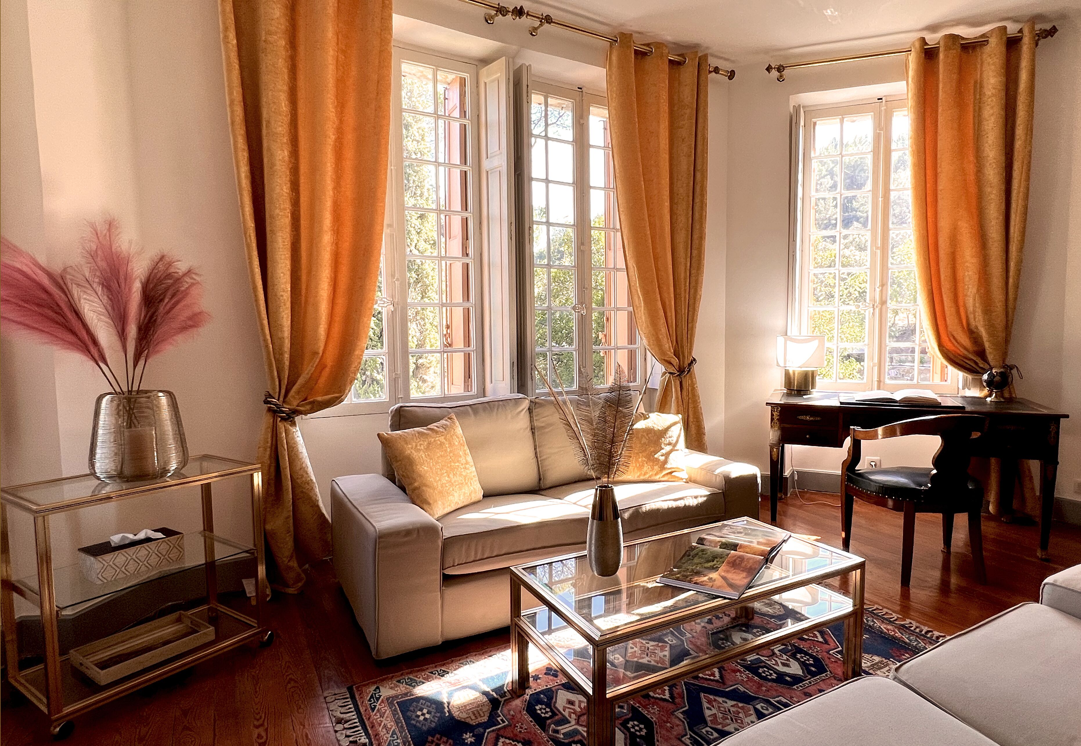 Un salon comprenant un canapé, une table basse en verre, un bureau et une chaise. Les fenêtres sont dotées de rideaux, et une table d’appoint est ornée d’un vase contenant des plantes séchées.