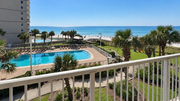 A pool with a view of the beach and ocean, surrounded by palm trees and a white fence.
