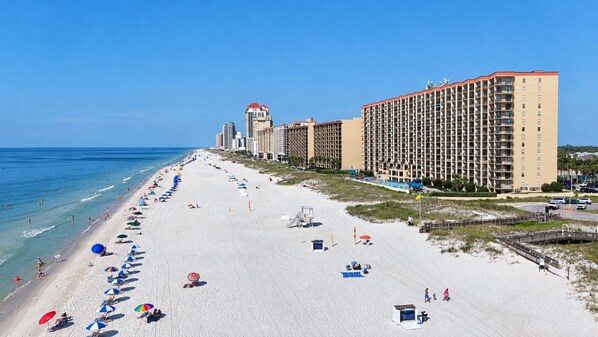 A beach with a row of beach umbrellas, a lifeguard tower, and a large hotel building with multiple floors. - Seaside Orange Beach, Indoor Pool, Tennis Courts! (Orange Beach)