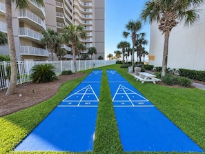 A shuffleboard court with numbered scoring areas, surrounded by a well-maintained lawn, palm trees, and a white fence.