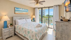 A bedroom with a bed, bedside lamps, a ceiling fan, a TV, and a view of the ocean through a balcony.