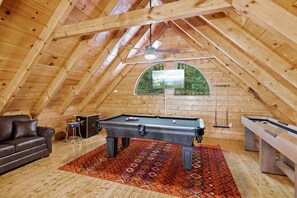 A cozy attic room with a pool table, a sofa, and a window overlooking greenery.