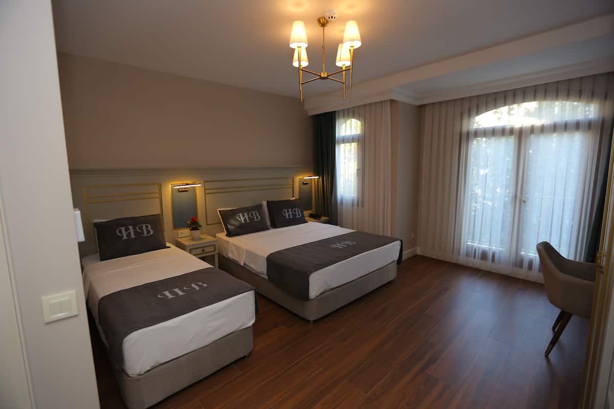 A hotel room with two beds, a chandelier, a nightstand, a chair, and a large window with curtains.