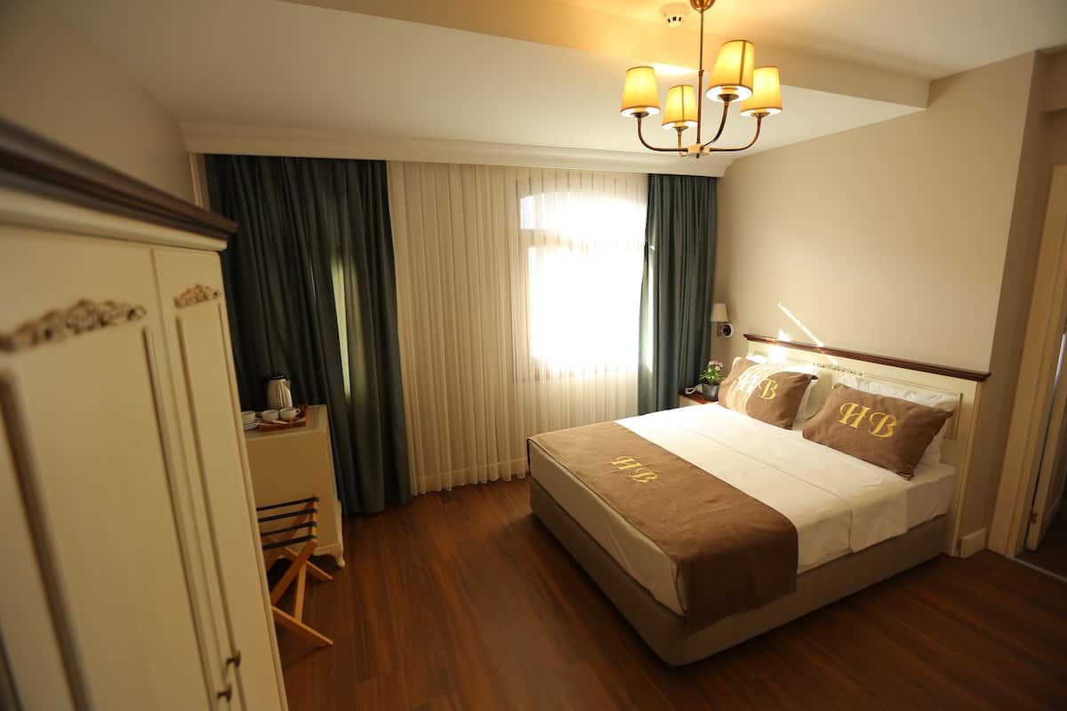 A hotel room with a large bed, a desk with a chair, a wardrobe, and a chandelier.