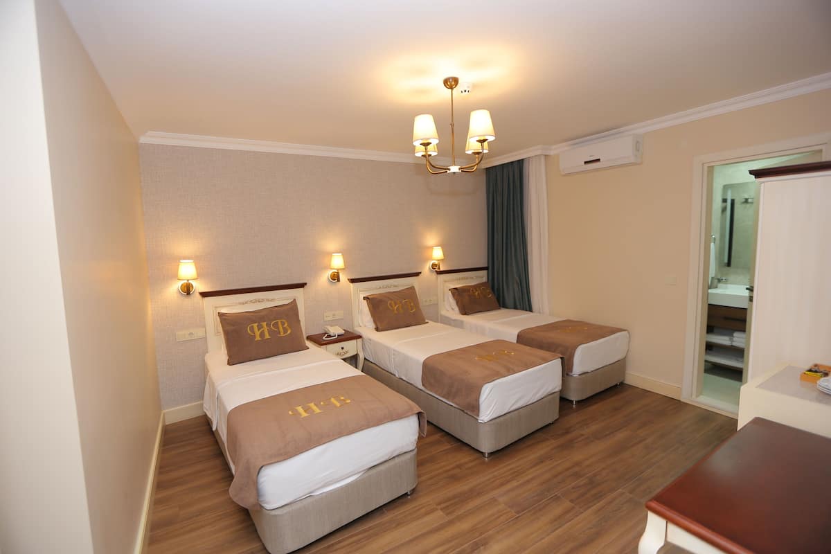A hotel room with three single beds, each with a headboard and a pillow with "H&B" embroidered on it. There is a wooden floor, a small table, and a ceiling light fixture.
