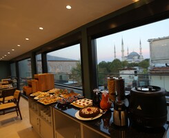 Un bufé de desayuno con una variedad de pasteles y bizcochos, una cafetera, y una vista a una mezquita con minaretes al fondo.