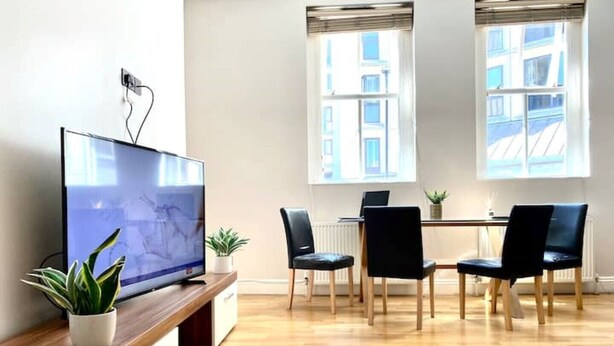 A modern living room with a flat-screen TV on a wooden stand, a dining table with four chairs, and two windows with blinds. - Sight Flat - SF01 - One Bedroom Flat (London)