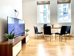 A modern living room with a flat-screen TV on a wooden stand, a dining table with four chairs, and two windows with blinds.