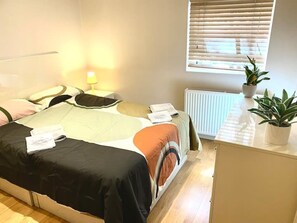 A neatly made bed with a variety of pillows and a blanket, a bedside table with a lamp, a radiator, and a window with blinds.