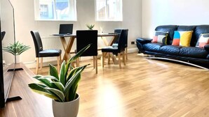 A modern living room with a black leather sofa, a dining table with chairs, and a large window providing natural light. - Sight Flat - SF01 - One Bedroom Flat (London)