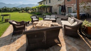 An outdoor patio with wicker furniture, a stone floor, and a view of a rustic building with a garden and mountains in the background. - Olimagio Farmstay w/animals & 25m pool <4km Forte dei Marmi beach (Pietrasanta)