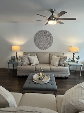 A living room with a beige sofa, a round wall decoration, a ceiling fan, and two lamps on side tables.