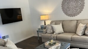 A living room with a sectional sofa, a coffee table, a TV, a lamp, and a decorative wall piece. - Cozy New Home, Close to Beaches, Restaurants and Bike Trials, Free EV charging  (Rehoboth Beach)
