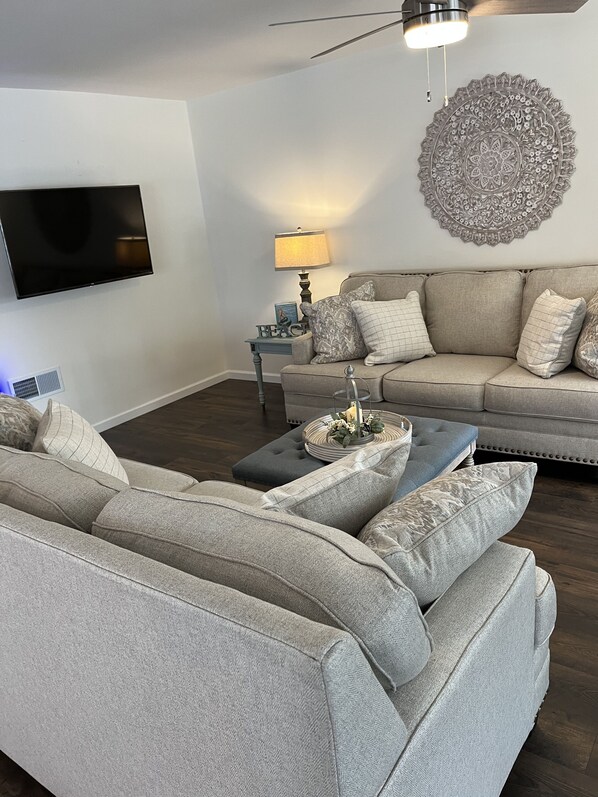 A living room with a sectional sofa, a coffee table, a TV, a lamp, and a decorative wall piece. - Cozy New Home, Close to Beaches, Restaurants and Bike Trials, Free EV charging (Rehoboth Beach)