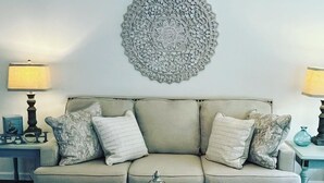 A living room with a beige sofa, two side tables with lamps, a decorative wall piece, and a tray with flowers on a matching ottoman. - Cozy New Home, Close to Beaches, Restaurants and Bike Trials, Free EV charging  (Rehoboth Beach)