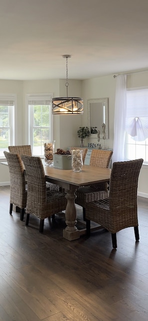 A dining room with a round table, wicker chairs, a hanging light fixture, and a mirror on the wall.