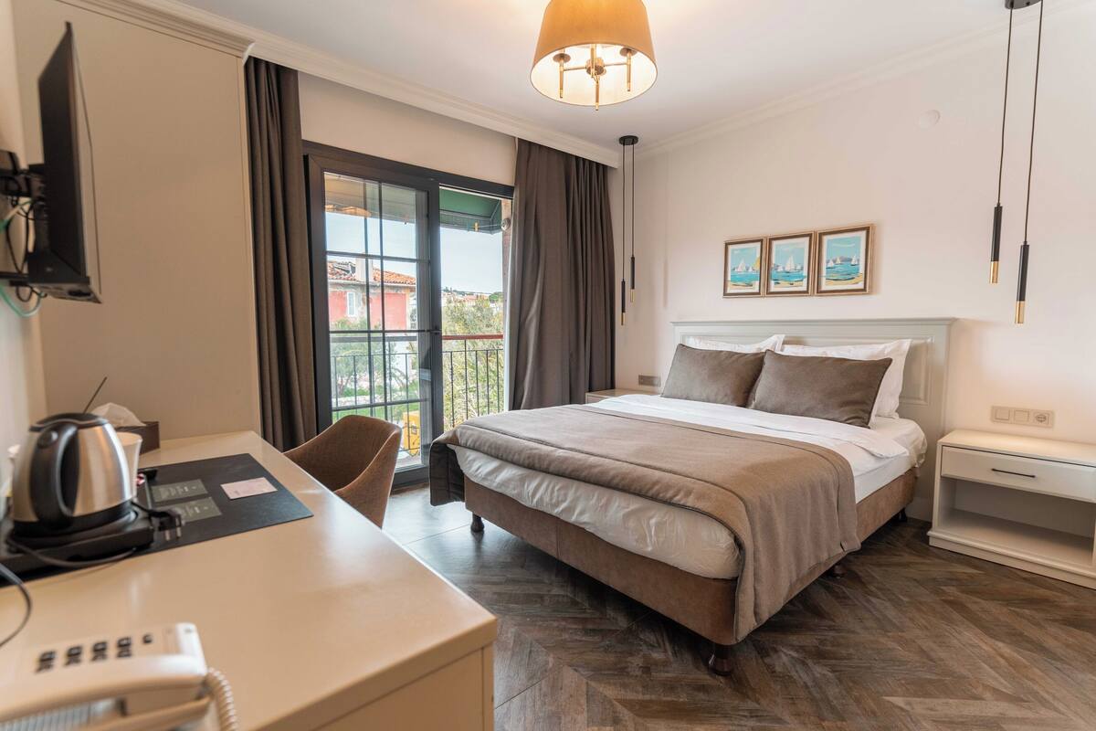 Hotelkamer met een bed, een bureau met telefoon, een stoel, een raam met gordijnen en een balkon.