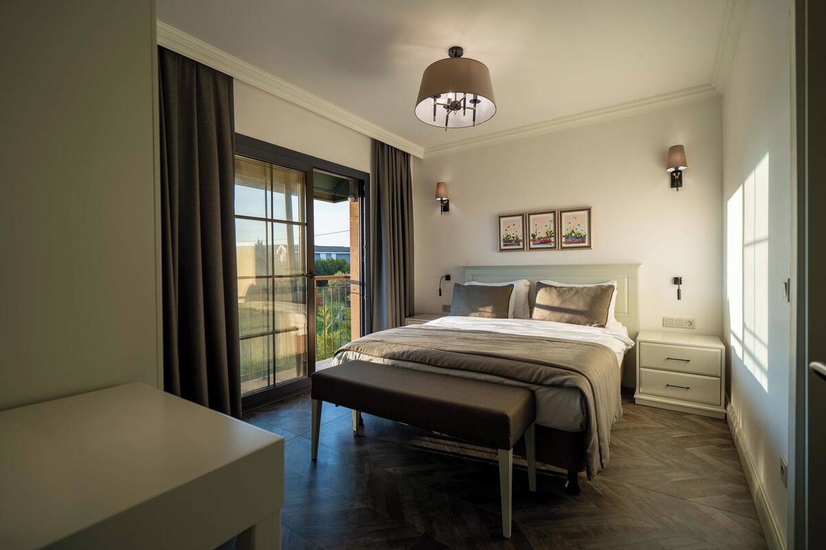 Een slaapkamer met een bed, een bankje, nachtkastjes, wandlampen en uitzicht door een groot raam.